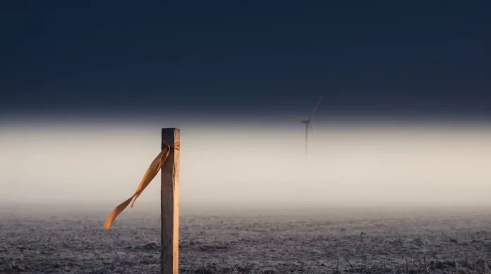 Windkraft-Schwindel – Nebelfeld mit Vermessungspflock Vermessungspflock mit orangefarbenem Markierungsband auf einem leeren Feld im dichten Nebel, dahinter die kaum sichtbare Silhouette eines Windrads – Sinnbild für verkaufte Windparks, die nie existierten