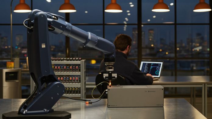 Robotikarm in einem Zürcher Labor zur blauen Stunde Navyblauer Roboterarm steckt an einer Werkbank einen Kabelstecker in ein Steuermodul, im Hintergrund ein Ingenieur am Laptop und die Zürcher Skyline zur blauen Stunde
